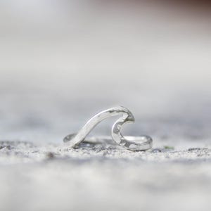 May include: A silver wave-shaped ring. The ring has a hammered texture and a curved design, resembling a breaking ocean wave. The ring is a piece of jewellery.