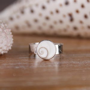 May include: A silver ring with a circular, white shell inlay displaying a spiral design. The ring sits on a wooden surface, with a blurred seashell in the background. The ring's band has a textured finish.