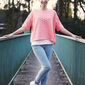 May include: A woman wearing a coral-colored top with a repeating pattern, light blue jeans, and white sneakers with pink accents. The top has short sleeves and a loose fit. The sneakers have a high-top design.