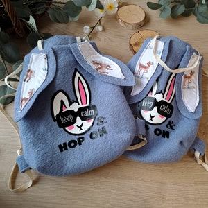 May include: Two blue felt bunny backpacks with white ears and a white bunny face with black eyes and a pink nose. The bunny has black sunglasses that say "keep calm". The backpacks have white fabric with a brown deer pattern on the ears. The backpacks say "Hop On" and "& On" in black text.