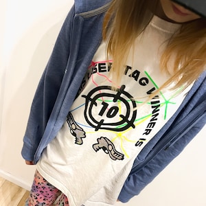 May include: White t-shirt with a black target graphic and the text "This Laser Tag Winner Is" in a colorful font. The shirt is being worn by a person with long blonde hair and a blue hoodie.