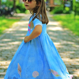 May include: A young girl wearing sunglasses and a light blue dress with a seashell design. The dress has a white trim and a pink lace hem. The girl is standing on a stone path.