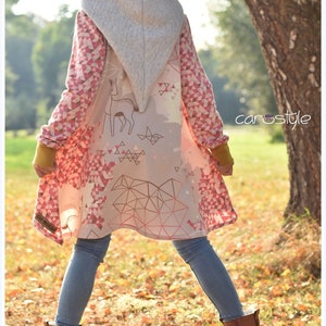 Puede incluir: Un abrigo largo con capucha con un estampado geométrico y de ciervos en rosa y blanco. El abrigo tiene una capucha gris y mangas con un estampado de triángulo rosa y blanco. La persona lleva vaqueros azules, botas marrones y un gorro rosa.