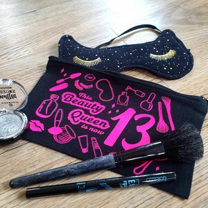 Può includere: Un beauty case nero con la scritta "This Beauty Queen is now 13" e illustrazioni rosa. Una mascherina per dormire blu navy con ciglia dorate, un pennello per trucco e un eyeliner sono visibili.