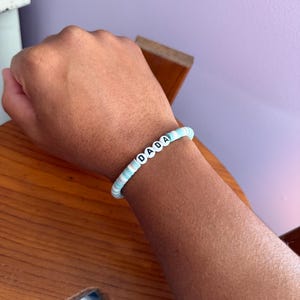 May include: A blue and white beaded bracelet spelling out the word "DADA" in white letter beads. The bracelet is worn on a wrist, with a wooden surface and a purple wall in the background.