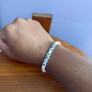 May include: A white beaded bracelet with the words "GIRL DAD" spelled out in black letters. The bracelet features alternating pink and white beads. The bracelet is worn on a wrist.