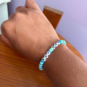 May include: A blue and white beaded bracelet with the word "BOY DAD" spelled out in white letter beads. The bracelet is on a wrist, with a wooden table in the background.