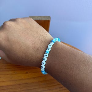 May include: A beaded bracelet with the words "GIRL DAD" spelled out in white letter beads. The bracelet features alternating blue and white beads. The bracelet is worn on a wrist.