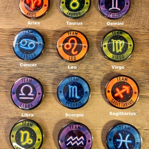 Puede incluir: Una colección de doce botones redondos, cada uno con un signo zodiacal diferente. Los botones tienen un acabado brillante y un borde negro. Cada botón tiene la palabra "TEAM" en la parte superior y el nombre del signo zodiacal en la parte inferior.