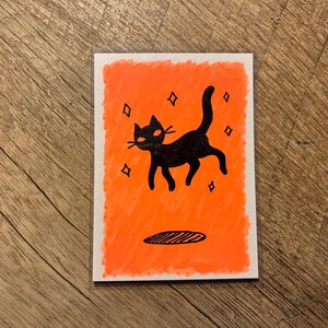 May include: A black cat illustration on a rectangular card with an orange background. The cat has a curved tail and is surrounded by diamond shapes. A black oval shape is at the bottom. The card is on a wooden surface.