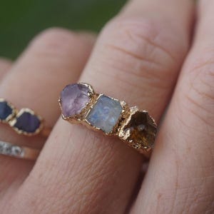 May include: A gold ring featuring three raw, uncut gemstones set in a textured metal band. The stones include a pale purple amethyst, a translucent moonstone with a blue sheen, and a warm brown citrine, all arranged horizontally across the finger.