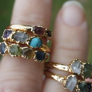 Peut inclure: Une collection de bagues dorées ornées de plusieurs pierres précieuses brutes et non taillées de différentes couleurs, notamment turquoise, transparent, vert et violet foncé. Les anneaux métalliques ont une finition texturée.