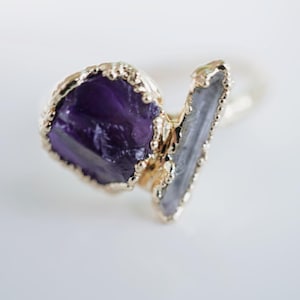 May include: A gold ring with a large purple amethyst stone and a smaller clear quartz stone. The stones are set in a gold bezel setting.