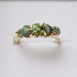May include: A gold ring featuring a row of five raw, unpolished green stones set along the top band. The metal band has a smooth, polished finish that contrasts with the textured, irregular surfaces of the natural green gemstones.
