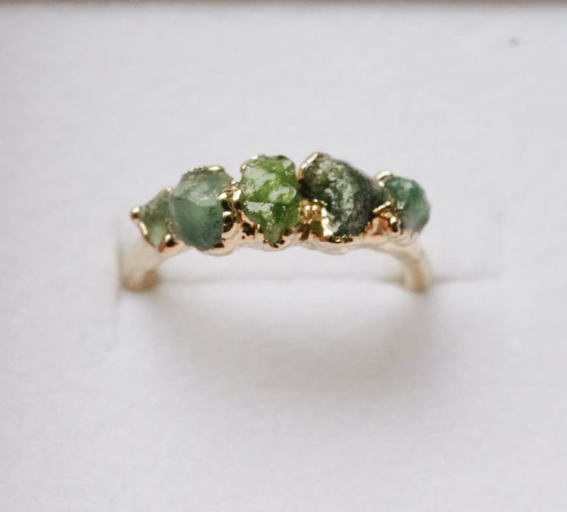 May include: A gold ring featuring a row of five raw, unpolished green stones set along the top band. The metal band has a smooth, polished finish that contrasts with the textured, irregular surfaces of the natural green gemstones.