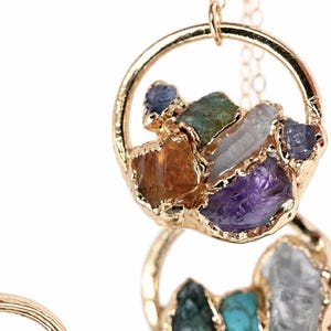 May include: A gold-toned necklace featuring circular pendants suspended from a delicate chain. Each pendant contains a cluster of raw, multi-colored gemstones including purple amethyst, amber citrine, blue sapphire, and clear quartz set in gold.