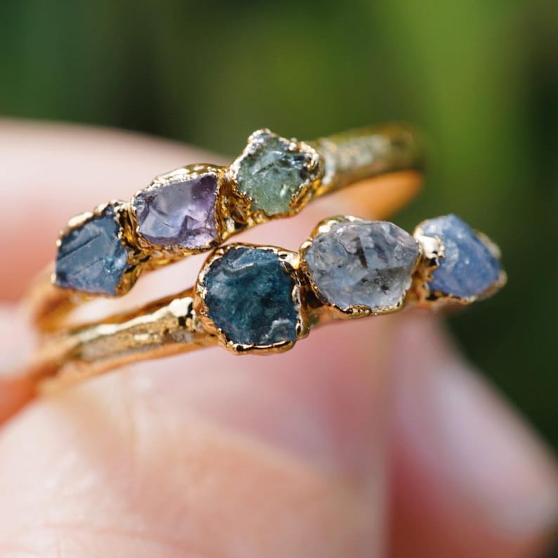 May include: Two gold rings featuring raw, uncut gemstones in various shades of blue, purple, and green. Each ring has a textured, organic metal band with multiple rough-cut stones set along the top, showcasing natural variations in color and shape.