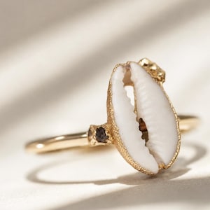 Könnte beinhalten: Ein goldener Ring mit einem zentralen weißen Kaurimuschel-Element mit einem texturierten, goldfarbenen Rand. Das Band ist dünn und poliert, mit einem kleinen, dunklen, rau texturierten Akzentstein auf jeder Seite der Muschel, was einen Kontrast erzeugt.