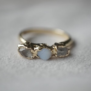 May include: A gold ring featuring a central round, opaque light blue stone flanked by two raw, translucent, irregular-shaped grey stones. The metal band has a textured, organic finish, and the stones are set in a prong-style setting.