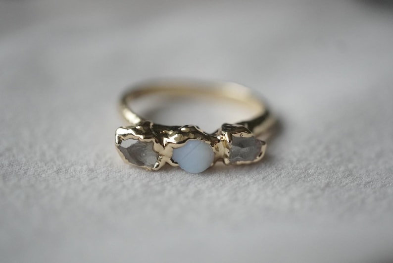 May include: A gold ring featuring a central round, opaque light blue stone flanked by two raw, translucent, irregular-shaped grey stones. The metal band has a textured, organic finish, and the stones are set in a prong-style setting.