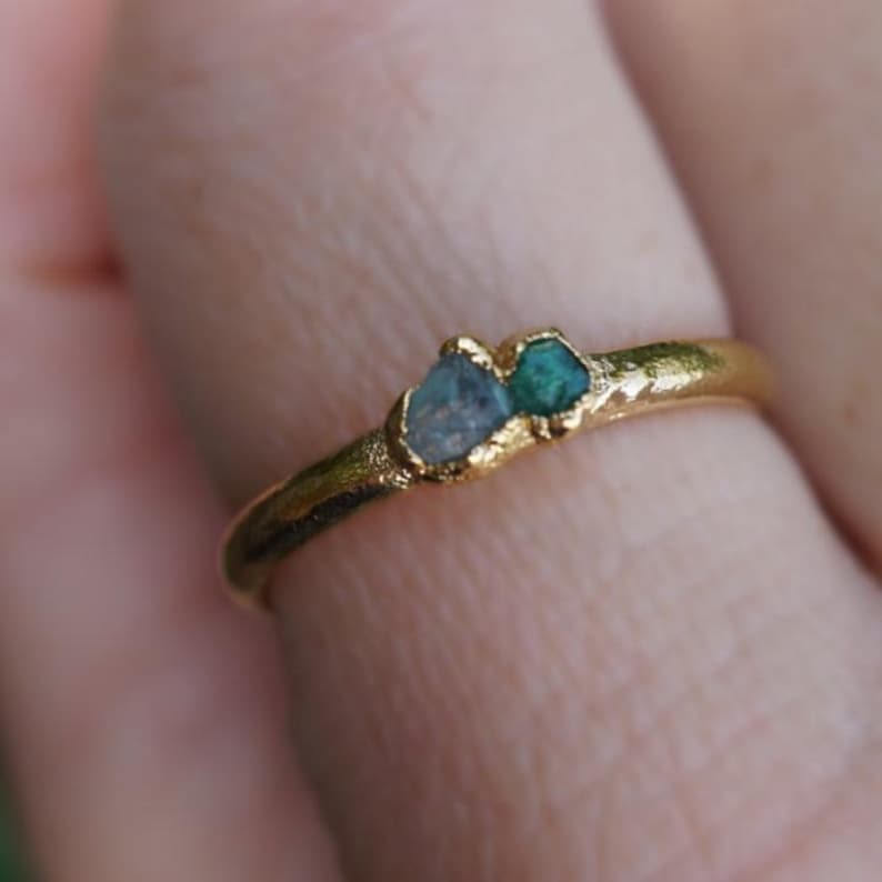 May include: A gold ring with a textured, organic band featuring two small, raw, uncut gemstones set side-by-side. One stone is a pale, translucent blue, and the other is a vibrant, deep green. The metal has a matte, hammered finish throughout.