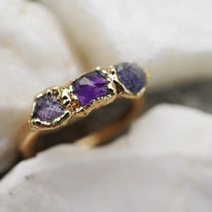 May include: A gold ring featuring three raw, uncut gemstones set in a textured gold band. The center stone is a deep purple amethyst, flanked by two smaller, lighter purple stones. The metal setting has a rough, organic finish around each stone.