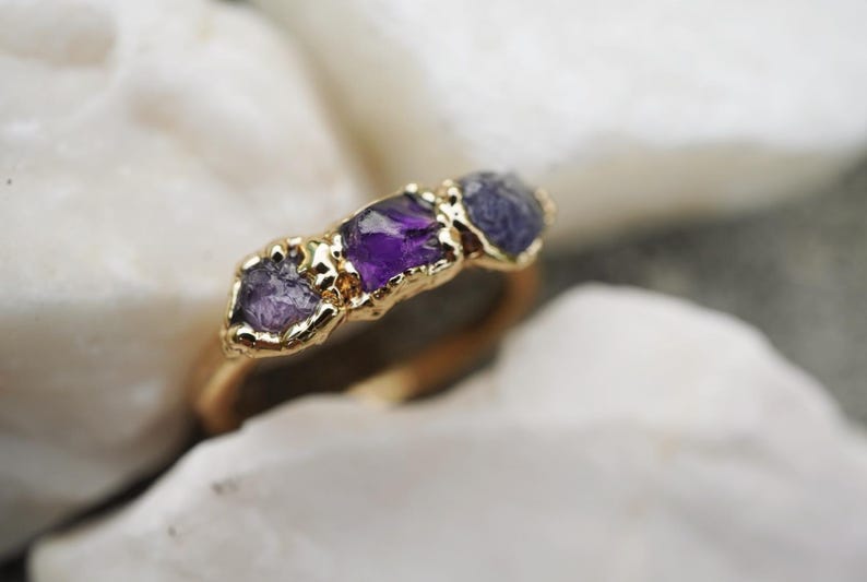May include: A gold ring featuring three raw, uncut gemstones set in a textured gold band. The center stone is a deep purple amethyst, flanked by two smaller, lighter purple stones. The metal setting has a rough, organic finish around each stone.