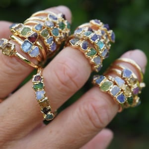 May include: A collection of gold-toned rings featuring multiple small, raw, multi-colored gemstones set along the bands. The stones display various hues including blue, green, yellow, and purple, with each ring showcasing a unique arrangement of these rough-cut mineral pieces.