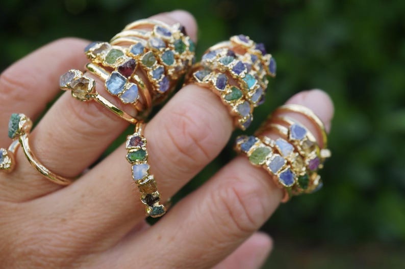 May include: A collection of gold-toned rings featuring multiple small, raw, multi-colored gemstones set along the bands. The stones display various hues including blue, green, yellow, and purple, with each ring showcasing a unique arrangement of these rough-cut mineral pieces.
