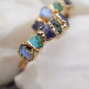 May include: A gold-toned ring featuring a series of raw, uncut gemstones in various colors including blue, turquoise, purple, and green. The stones are set in a textured gold metal band that follows a wavy, organic shape along the finger.