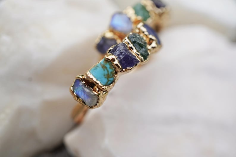 May include: A gold-toned ring featuring a series of raw, uncut gemstones in various colors including blue, turquoise, purple, and green. The stones are set in a textured gold metal band that follows a wavy, organic shape along the finger.