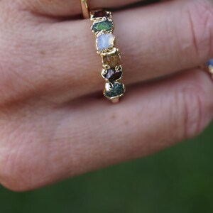 May include: A gold ring featuring a series of small, raw, multi-colored gemstones set in a vertical row. The stones include shades of deep red, green, white, and brown, each with a rough, natural texture held within a gold metal band.