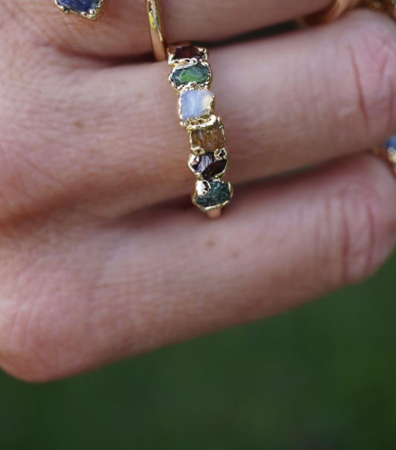 May include: A gold ring featuring a series of small, raw, multi-colored gemstones set in a vertical row. The stones include shades of deep red, green, white, and brown, each with a rough, natural texture held within a gold metal band.