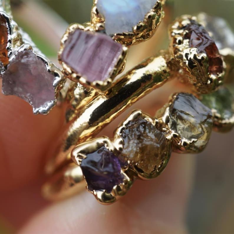 May include: A gold-toned ring featuring a series of raw, uncut gemstones in various colors including purple, amber, and brown, set in a textured metal band. The stones are irregularly shaped and held in place by small, jagged metal prongs.