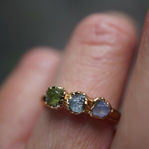 May include: A gold ring featuring three small, raw, uncut gemstones set in a row. The stones are arranged by color, displaying a deep olive green, a muted blue-grey, and a pale lavender-blue hue. The metal band has a polished gold finish.
