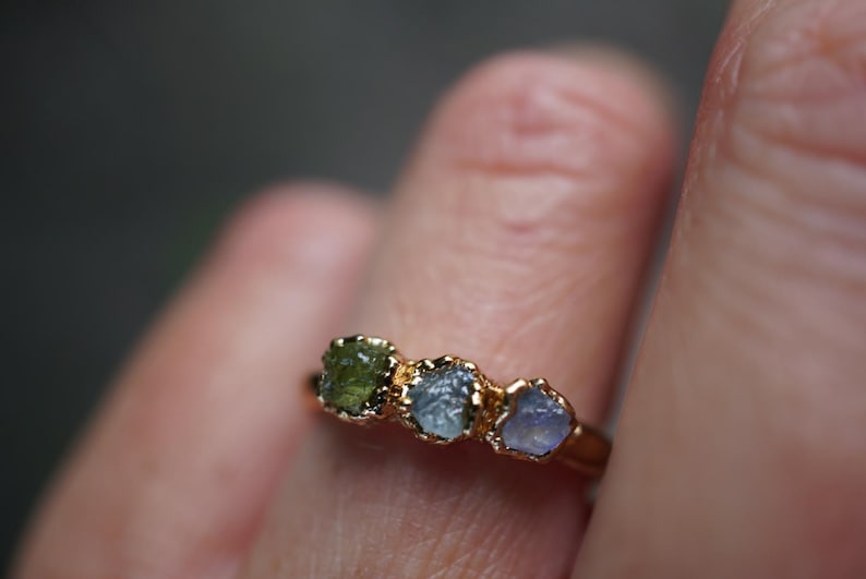 May include: A gold ring featuring three small, raw, uncut gemstones set in a row. The stones are arranged by color, displaying a deep olive green, a muted blue-grey, and a pale lavender-blue hue. The metal band has a polished gold finish.