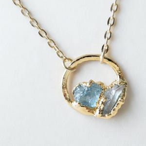 May include: A gold necklace featuring a circular pendant with a textured, hammered finish. The pendant holds two raw, unpolished gemstones: one light blue aquamarine and one clear, translucent crystal, both set within the gold circle frame.