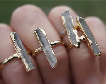 Könnte beinhalten: Nahaufnahme von drei goldfarbenen Ringen, jeder mit einem rechteckigen, grauen Stein mit goldfarbenem Rand. Die Ringe werden an Fingern getragen, um das Design zu zeigen. Der Hintergrund ist unscharf.