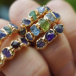 May include: A collection of gold-toned rings featuring multiple raw, uncut gemstones in various colors including blue, green, purple, and yellow. Each ring band is textured and holds several small, irregular, rough-cut stones set in a row.