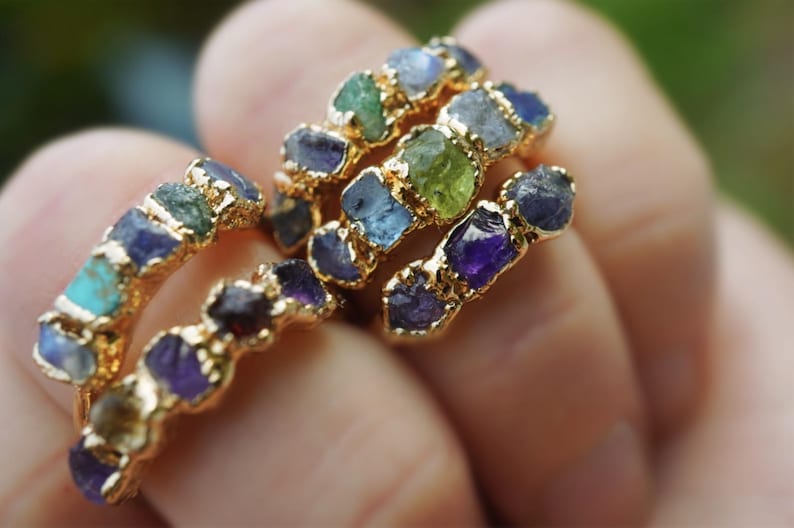May include: A collection of gold-toned rings featuring multiple raw, uncut gemstones in various colors including blue, green, purple, and yellow. Each ring band is textured and holds several small, irregular, rough-cut stones set in a row.