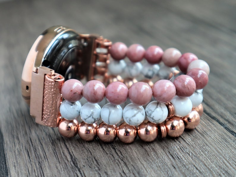 May include: A rose gold smartwatch with a beaded band. The band is made of three rows of beads: a row of white beads, a row of pink beads, and a row of copper beads.