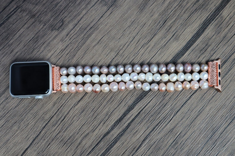 Puede incluir: Una pulsera de Apple Watch de oro rosa con un dise&ntilde;o de perlas de tres hilos. Las perlas son un degradado de rosa, blanco y crema.