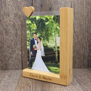 May include: A wooden photo frame with a heart-shaped accent holding a wedding photo. The frame is made of light-colored wood and features the names "Karen & Aaron" engraved on the base. The photo shows a bride and groom.