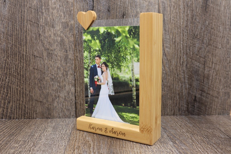 May include: A wooden photo frame with a heart-shaped accent holding a wedding photo. The frame is made of light-colored wood and features the names "Karen & Aaron" engraved on the base. The photo shows a bride and groom.
