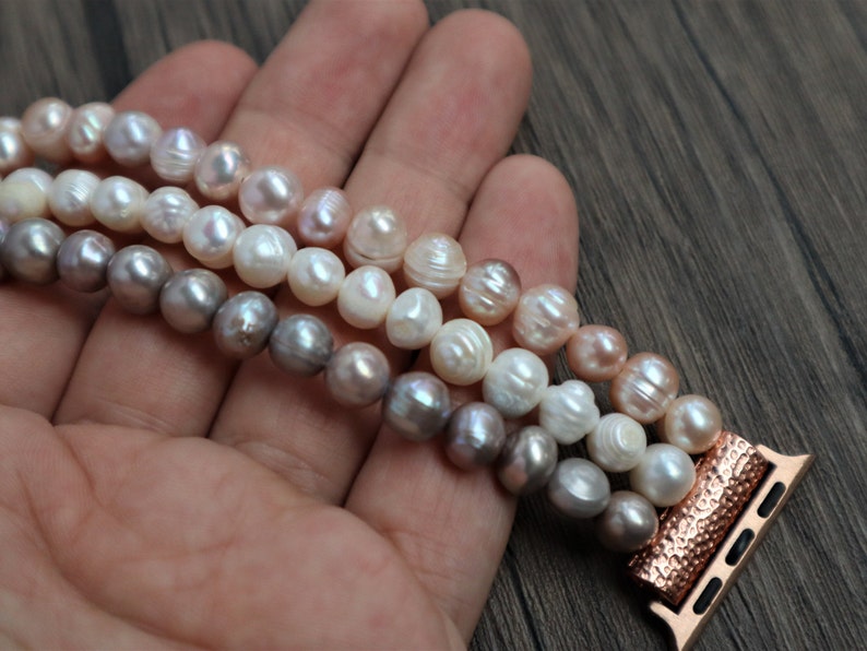 Puede incluir: Un brazalete de perlas de tres hilos con un adaptador de banda de Apple Watch de oro rosa. Las perlas son en tonos de blanco, rosa y gris.