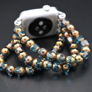 May include: A watch band made of blue and gold beads. The band is made of two rows of beads, with the blue beads on the outside and the gold beads on the inside. The band is wrapped around a silver Apple Watch.