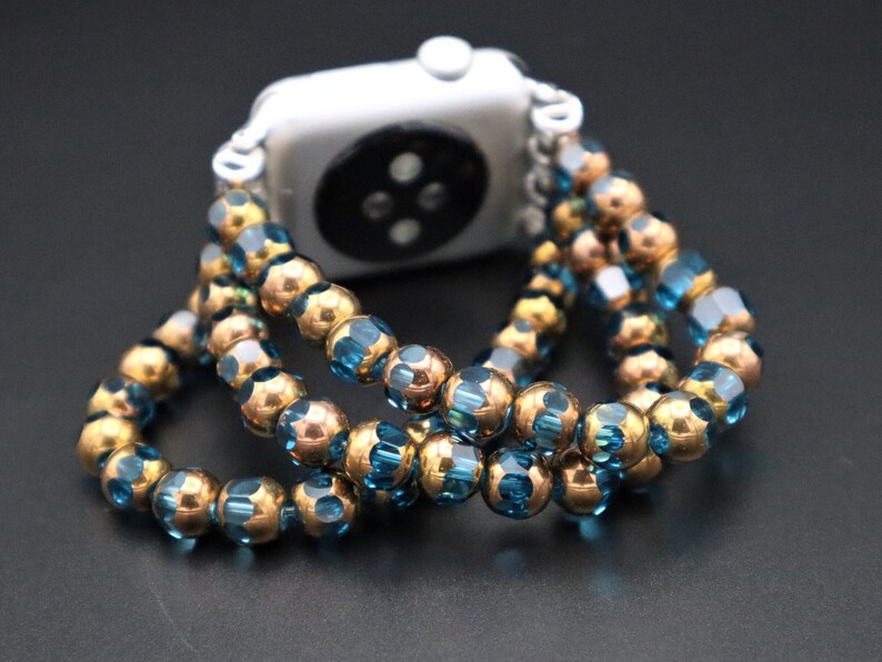 May include: A watch band made of blue and gold beads. The band is made of two rows of beads, with the blue beads on the outside and the gold beads on the inside. The band is wrapped around a silver Apple Watch.