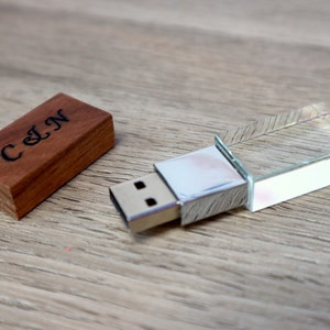 K&ouml;nnte beinhalten: Ein USB-Stick aus Holz mit den Initialen "C & N" auf der Holzabdeckung eingraviert. Der USB-Stick hat ein klares, geometrisches Kristall-Design. Die Artikel werden auf einer hellbraunen Holzoberfl&auml;che pr&auml;sentiert.