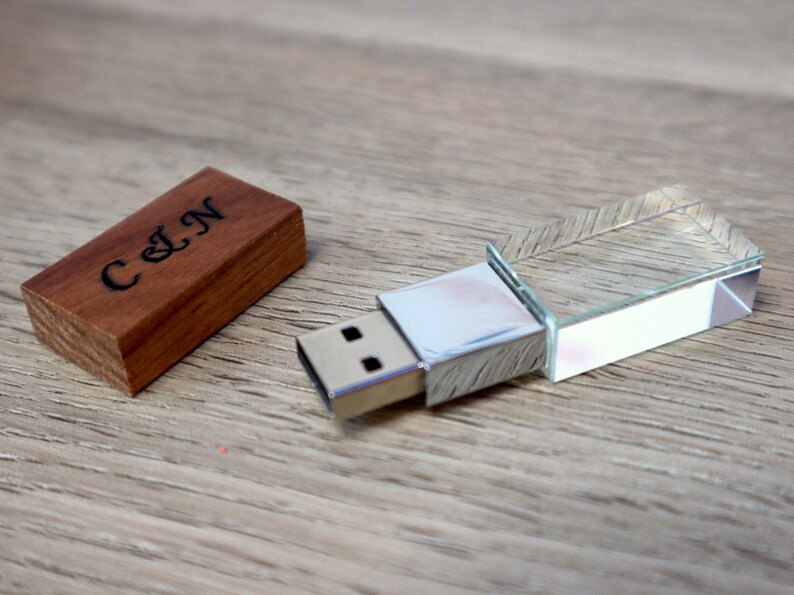K&ouml;nnte beinhalten: Ein USB-Stick aus Holz mit den Initialen "C & N" auf der Holzabdeckung eingraviert. Der USB-Stick hat ein klares, geometrisches Kristall-Design. Die Artikel werden auf einer hellbraunen Holzoberfl&auml;che pr&auml;sentiert.