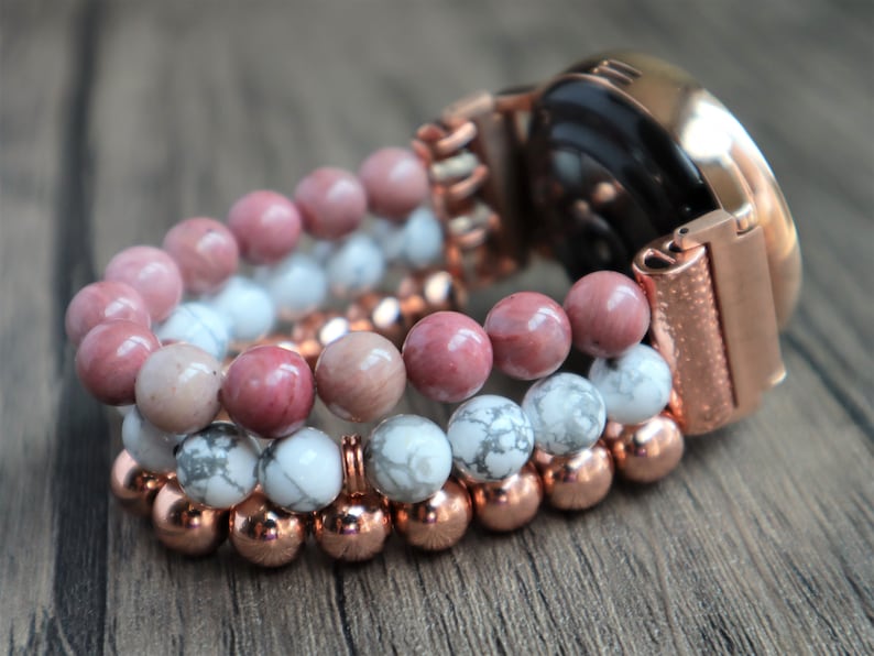 May include: A rose gold smartwatch with a beaded band. The band is made of three strands of beads: pink, white, and copper.